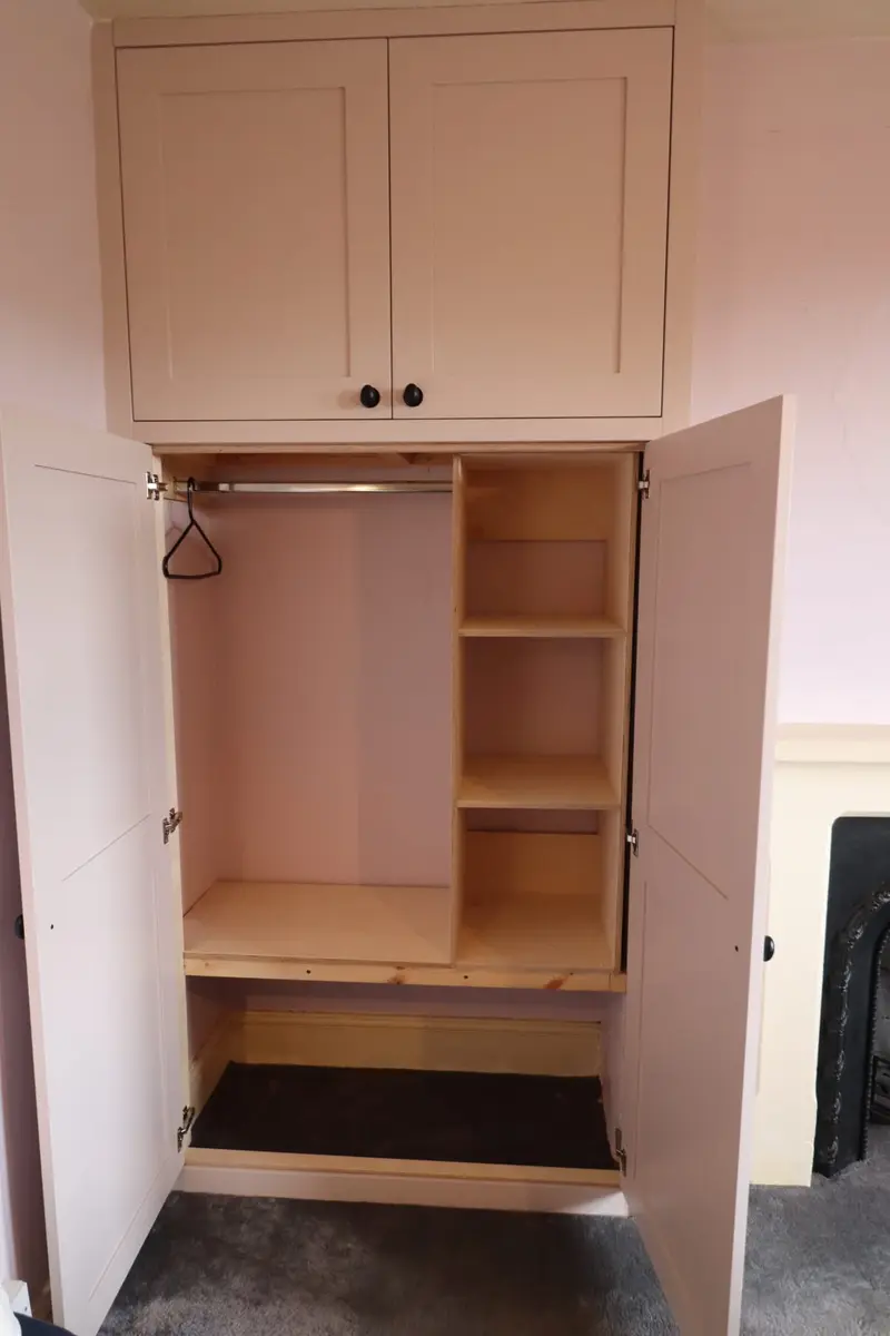 Alcove wardrobe painted dusty pink, doors open on a hanging rail and birch ply shelves, with a storage cupboard above