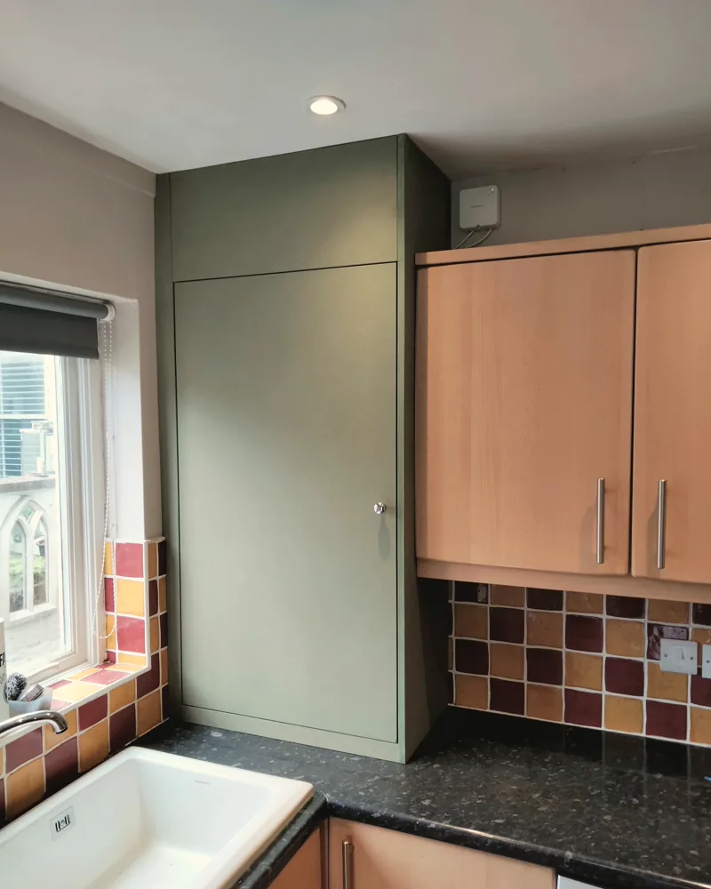 Sage green cupboard built to enclose a kitchen boiler, floor to ceiling beside the window